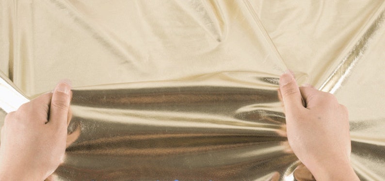 Op de afbeelding: Een gouden metallic stof wordt vastgehouden door twee handen. De stof is glad en glanzend, met een lichte glans.