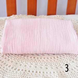 Puede incluir: Una manta de muselina rosa p&aacute;lido arrugada. La manta est&aacute; doblada y colocada sobre una superficie de crochet blanco.