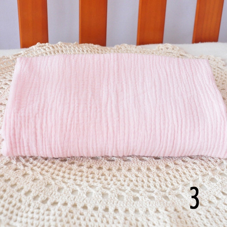 Puede incluir: Una manta de muselina rosa p&aacute;lido arrugada. La manta est&aacute; doblada y colocada sobre una superficie de crochet blanco.