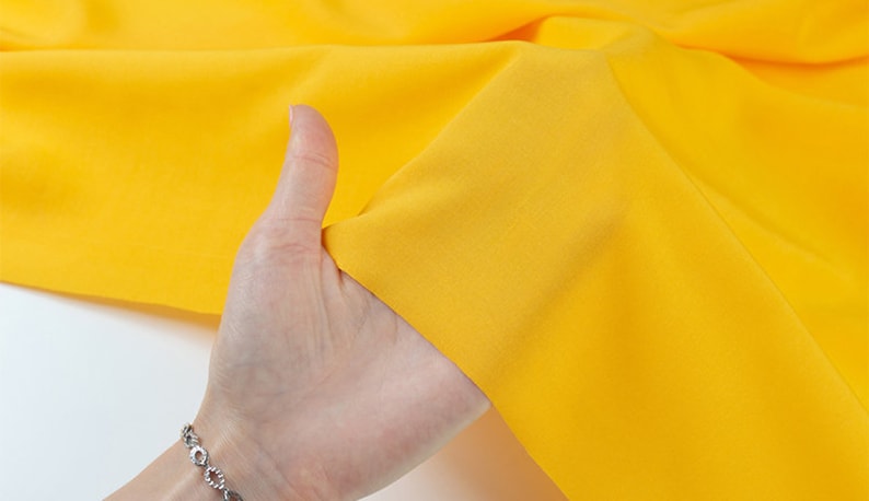 K&ouml;nnte beinhalten: Ein leuchtend gelber Stoff mit glatter Textur. Der Stoff wird von einer Hand gehalten.