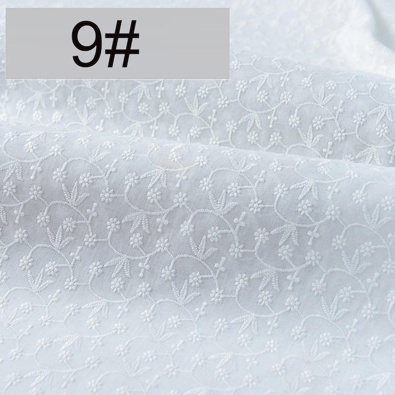 May include: A white fabric featuring an intricate, repeating embroidered pattern of small flowers and delicate leaves. The textile has a soft, folded texture and includes a rectangular overlay in the top left corner displaying the text 9#.