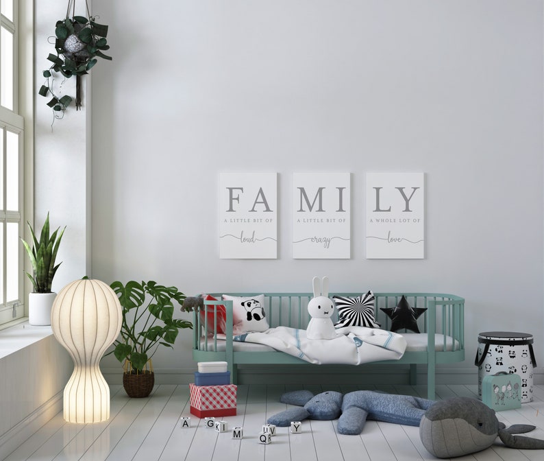 K&ouml;nnte beinhalten: Ein hellblaues Kinderbett mit einer wei&szlig;en Decke und einem Stoffhasen. An der Wand hinter dem Bett h&auml;ngen drei Leinw&auml;nde mit dem Wort "FAMILY" und einem Spruch &uuml;ber Liebe, Verr&uuml;cktheit und Last.