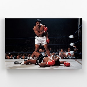 May include: A black and white photograph of two boxers in a boxing ring. The boxer on the left is lying on the ground, while the boxer on the right is standing over him with his right foot on the fallen boxer's chest. The boxer on the right is wearing white shorts and red boxing gloves. The boxer on the left is wearing black shorts and red boxing gloves. The photograph is taken from a low angle, looking up at the boxers.