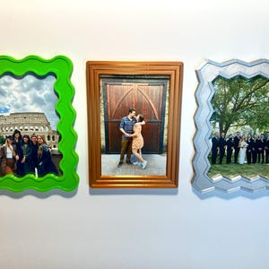 May include: Three picture frames with different coloured frames and different photos inside. The frame on the left is green with a wavy edge and a photo of a group of people in front of the Colosseum in Rome. The middle frame is brown with a photo of a couple kissing in front of a wooden door. The frame on the right is silver with a wavy edge and a photo of a wedding party standing in front of a tree.