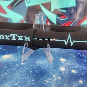 Puede incluir: Correa de reloj negra con el texto "VoxTek" en azul claro, junto con un dise&ntilde;o de l&iacute;nea de latido card&iacute;aco azul claro. La correa se muestra sobre un soporte transparente contra un fondo azul y blanco con tem&aacute;tica de galaxia.