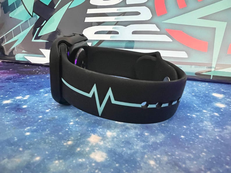 Puede incluir: Reloj inteligente negro con un dise&ntilde;o de latido card&iacute;aco azul claro en la correa. El reloj est&aacute; sobre un fondo con un dise&ntilde;o espacial. La correa tiene una serie de peque&ntilde;os agujeros.