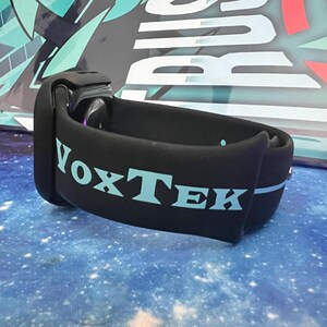 Puede incluir: Brazalete negro con la palabra "VoxTek" en letras azul claro. La pulsera es curva y parece estar hecha de un material suave y flexible. El fondo es una superficie azul con un patr&oacute;n de galaxia.
