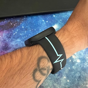 Puede incluir: Correa de reloj inteligente negra con un dise&ntilde;o de latido card&iacute;aco azul claro. La correa est&aacute; envuelta alrededor de una mu&ntilde;eca, mostrando el detalle del dise&ntilde;o. Un accesorio elegante.