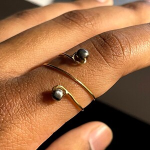 May include: A gold-colored, wrap-around ring with two dark gray, spherical accents. The ring is worn on a finger, with the band spiraling around the finger. The ring's design is minimalist and modern, with a focus on the contrast between the gold and gray.