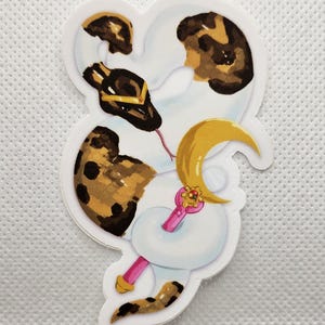 May include: A die-cut sticker featuring a cartoon snake with a white body, brown spots, and a gold crescent moon. The snake has a pink tongue and a gold crown. The sticker has a white border.