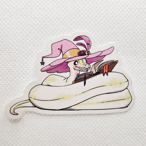 May include: A sticker featuring a cartoon snake wearing a pink witch hat with a feather and star charm, reading a book. The snake is white with yellow accents, and the book has a red bookmark.