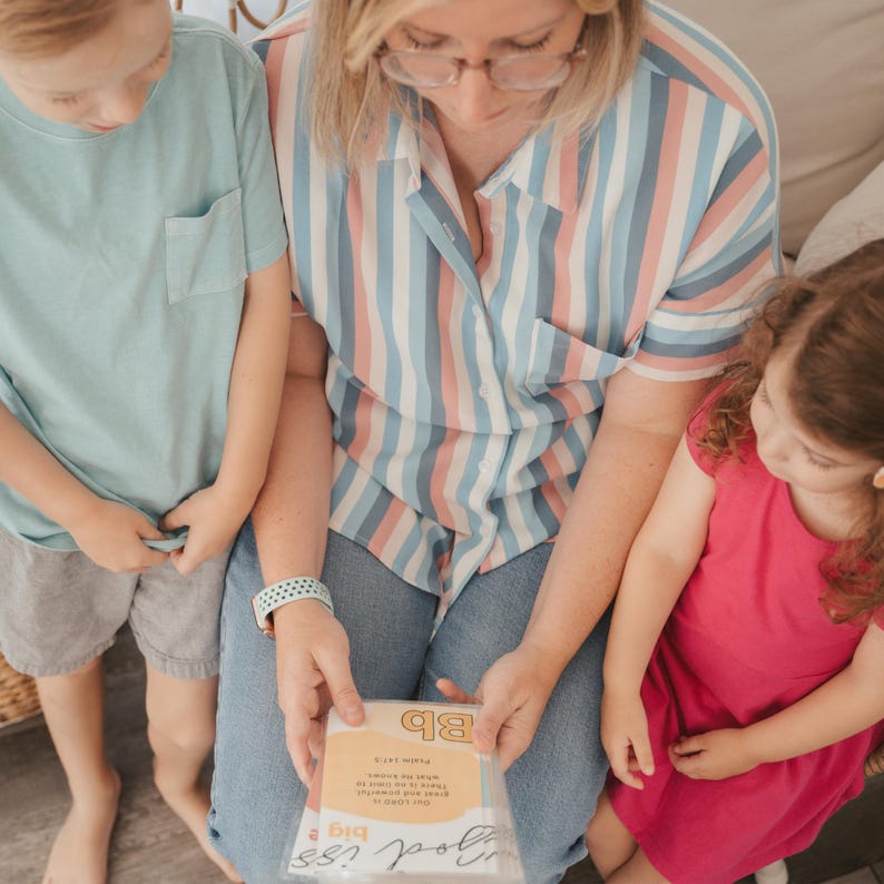 May include: A woman and two children are looking at a card that says "Good Job! You're a Big Deal!" The card is yellow with blue and orange accents. The woman is wearing a blue and pink striped shirt and jeans. The children are wearing blue and pink shirts.
