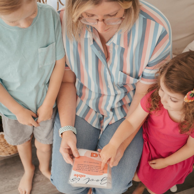 May include: A woman and two children are looking at a small card that says "God is able" in a pink and white striped shirt, blue jeans, and a pink shirt.