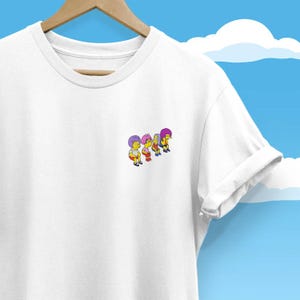 May include: White t-shirt featuring a small, colourful graphic of four cartoon characters. The characters have different coloured hair and clothing. The t-shirt is hanging on a wooden hanger against a blue sky background with white clouds.