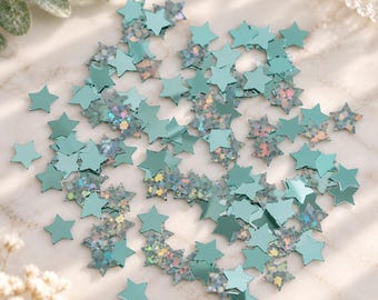 May include: A collection of small, five-pointed star-shaped confetti pieces in a mix of solid teal and holographic silver finishes. The stars are scattered across the surface, displaying a reflective, metallic appearance with varying light-catching textures.