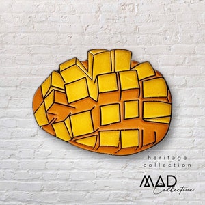 Puede incluir: Un pin de esmalte amarillo y naranja con forma de mango. El pin está formado por pequeños cuadrados que se asemejan a los segmentos de un mango. El pin tiene un contorno negro y está sobre un fondo de ladrillo blanco. El texto "heritage collection MAD Collective" es visible debajo del pin.