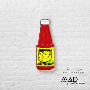 Puede incluir: Un pin de esmalte rojo y dorado con forma de botella de ketchup con las palabras "Banana Ketchup" impresas en la etiqueta. La etiqueta presenta un plátano amarillo con hojas verdes.