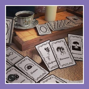 Puede incluir: Un juego de cartas de oráculo en blanco y negro con varios símbolos y texto en francés. Las cartas están dispuestas sobre una mesa de madera con una taza de café y una vela en el fondo.