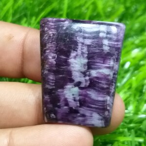 Peut inclure: Une pierre précieuse cabochon de couleur violette et blanche avec un motif strié. La pierre précieuse est taillée en forme rectangulaire et a une surface lisse et polie.