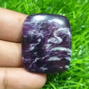 Peut inclure: Une pierre précieuse rectangulaire polie, de couleur violet foncé avec des motifs tourbillonnants blancs et vert pâle. La pierre est présentée sur un fond d'herbe verte, mettant en valeur ses veines naturelles.