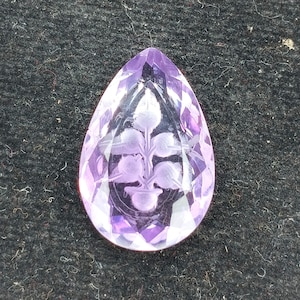 Puede incluir: Un colgante de amatista púrpura con un diseño floral tallado. El colgante está facetado y tiene forma de lágrima.