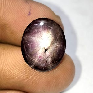 Könnte beinhalten: Ein oval geschliffener Edelstein in tiefem Lila, der einen Sterneneffekt aufweist. Der Stein hat eine glänzende Oberfläche mit inneren Linien und einem zentralen dunklen Punkt. Der Edelstein wird zwischen den Fingern gehalten und zeigt seine Größe und Farbe.