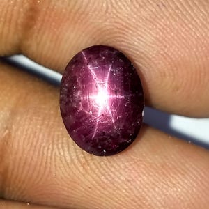 May include: Oval-shaped, deep red gemstone with a six-rayed star effect. The polished surface reflects light, creating a bright star pattern across the stone. The gem is held in a hand.