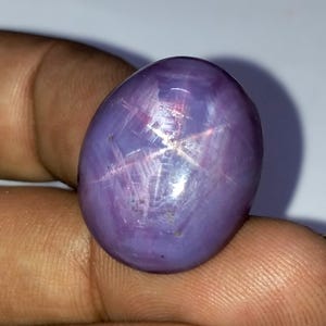 Puede incluir: Una gema pulida, de forma ovalada, de color púrpura, con un patrón de estrella de seis rayos. El efecto de estrella, o asterismo, se crea por la luz que se refleja en las inclusiones dentro de la piedra. La gema se sostiene en una mano.