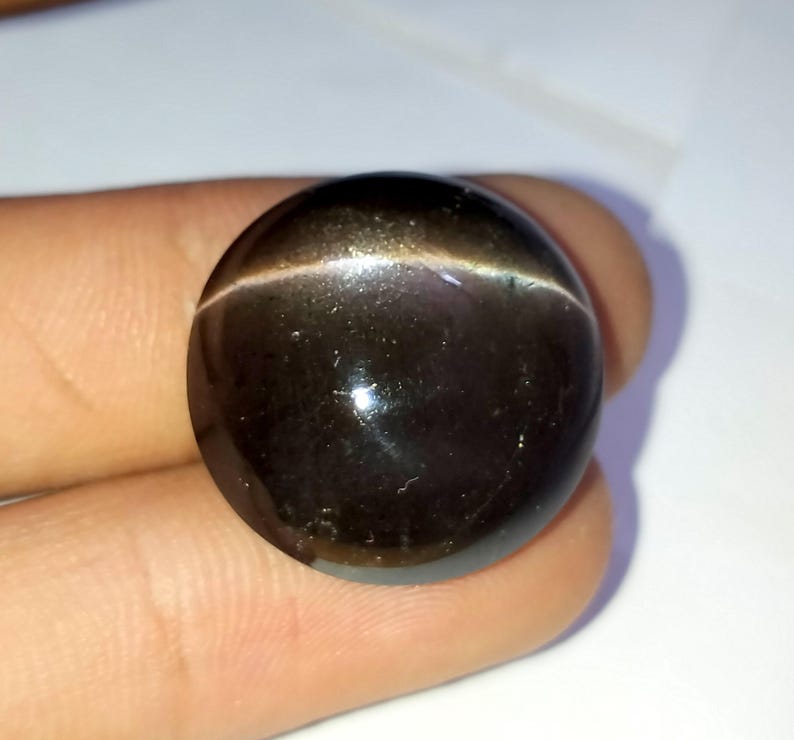 May include: A round, black gemstone with a golden sheen. The gemstone has a cat's eye effect, with a thin line of light running through the center.
