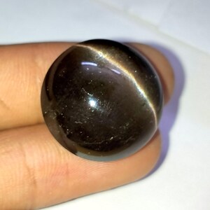 May include: A round, dark gray gemstone with a shimmering, iridescent effect. The gemstone has a smooth, polished surface.