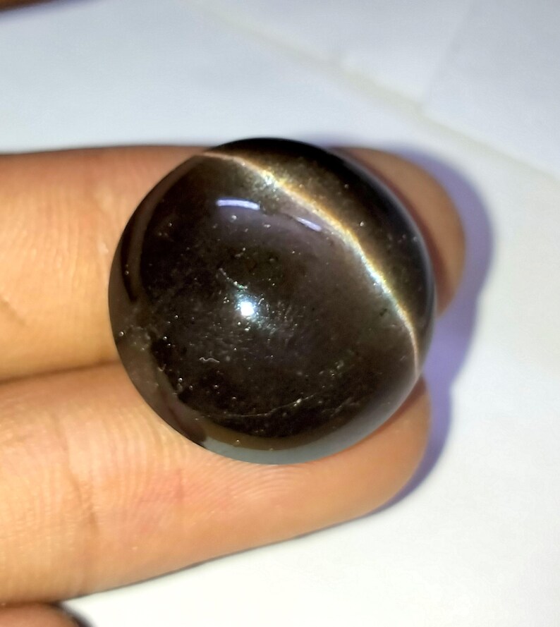 May include: A round, dark gray gemstone with a shimmering, iridescent effect. The gemstone has a smooth, polished surface.