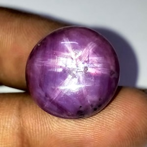 May include: A polished, round, purplish-red gemstone with a six-rayed star pattern. The star effect is created by light reflecting off of inclusions within the stone. The gem is held in a person's hand, showcasing its color and luster.