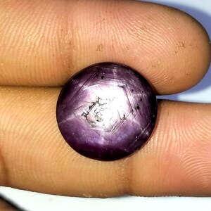 Könnte beinhalten: Ein runder, polierter, purpurroter Edelstein mit sternförmigem Muster. Der Edelstein hat eine glatte, reflektierende Oberfläche mit einem zentralen Stern und dunklen Einschlüssen. Der Edelstein wird zwischen Fingern gehalten.