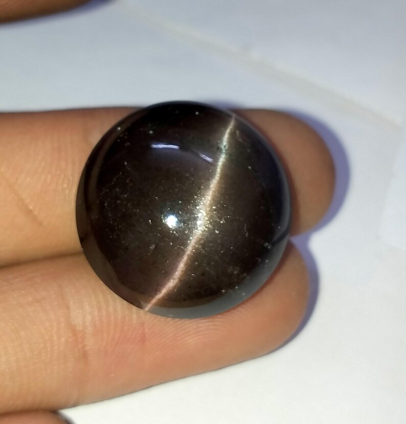 May include: A dark gray oval cabochon gemstone with a silver sheen. The gemstone has a cat's eye effect, with a single line of light running through the center.