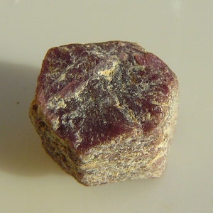 May include: A rough, dark red ruby gemstone with a hexagonal shape. The ruby has a natural, unpolished finish.