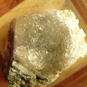 Può includere: Primo piano di un campione minerale cristallino di colore grigio chiaro con una superficie ruvida e irregolare. Il minerale è circondato da un materiale di aspetto metallico grigio scuro.