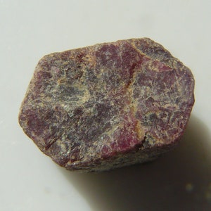 May include: A rough, dark red ruby gemstone with a slightly irregular shape.