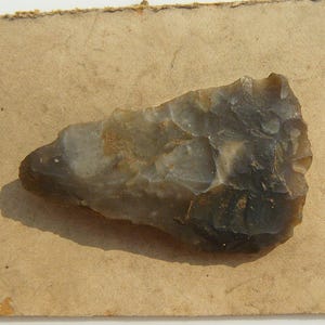 May include: A grey and brown stone tool, possibly a prehistoric hand axe, sits on a piece of aged, tan paper. The stone has a rough, textured surface with a dark grey edge and lighter grey and brown patches. The paper has a torn edge.