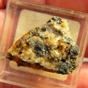 Op de afbeelding: Een klein, driehoekig gesteentemonster met een mix van licht- en donkergrijze kleuren. De steen heeft een ruw, getextureerd oppervlak en lijkt uit meerdere mineralen te bestaan.