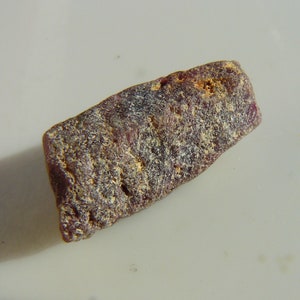 Puede incluir: Una piedra preciosa rugosa de color púrpura oscuro y gris, con una forma ligeramente rectangular.