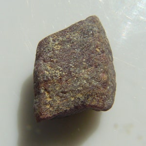 Könnte beinhalten: Ein rauer, dunkelroter Edelstein mit einer leicht unregelmäßigen Form. Der Edelstein hat eine matte, stumpfe Oberfläche und scheint ungeschliffen zu sein.