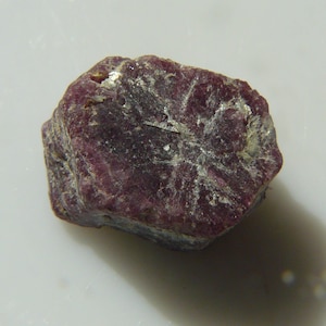 May include: A rough, dark red ruby gemstone with a natural, unpolished finish.