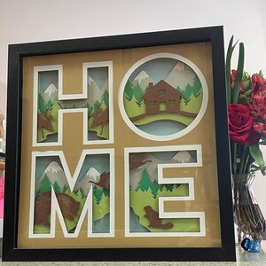 Peut inclure: Une œuvre d'art en papier encadrée avec le mot "HOME" en lettres blanches. Chaque lettre présente un design en papier superposé représentant une scène de montagne avec des arbres, des animaux et une petite maison.