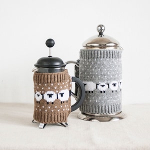 Könnte beinhalten: Zwei French Press Kaffeebereiter mit Strickhüllen. Einer ist braun mit weißen Schafen, der andere grau mit weißen Schafen. Beide haben schwarze Griffe, auf einer weißen Oberfläche.