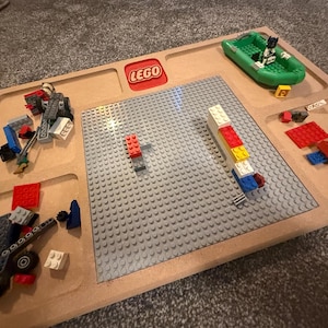 May include: A wooden LEGO play tray with a gray baseplate and various LEGO bricks and figures. A green boat with a LEGO figure is in the upper right corner. The LEGO logo is in red.