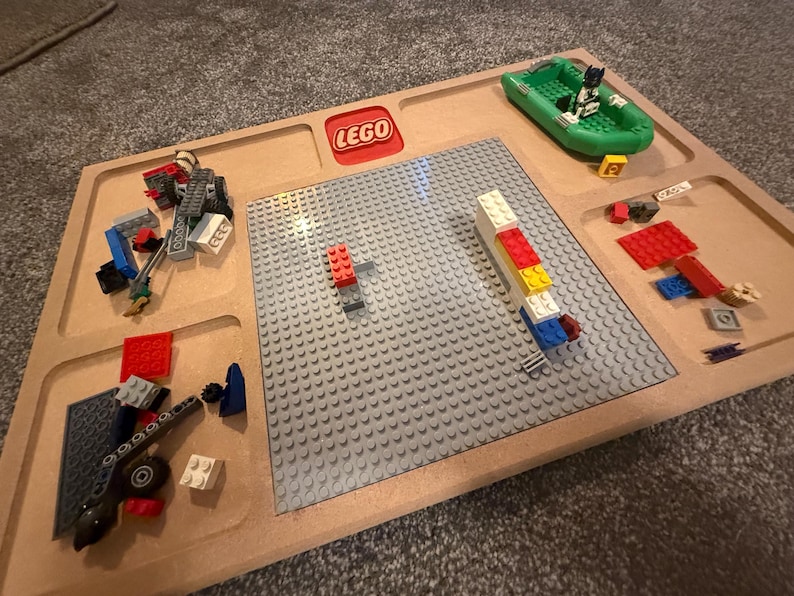 May include: A wooden LEGO play tray with a gray baseplate and various LEGO bricks and figures. A green boat with a LEGO figure is in the upper right corner. The LEGO logo is in red.