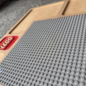 May include: A wooden LEGO baseplate with a gray surface for building. The LEGO logo is visible in a red rectangle. The baseplate has a raised edge and is designed to hold LEGO bricks.