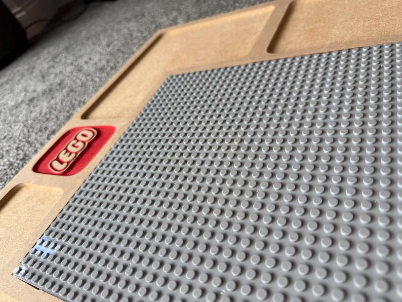 May include: A wooden LEGO baseplate with a gray surface for building. The LEGO logo is visible in a red rectangle. The baseplate has a raised edge and is designed to hold LEGO bricks.