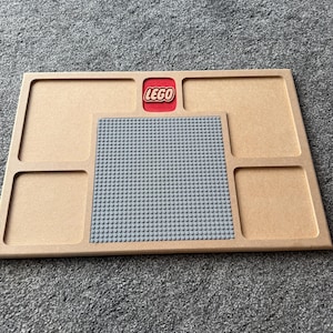 May include: A wooden Lego baseplate with a gray Lego building surface and four compartments. The Lego logo is in a red square at the top. The baseplate is tan.