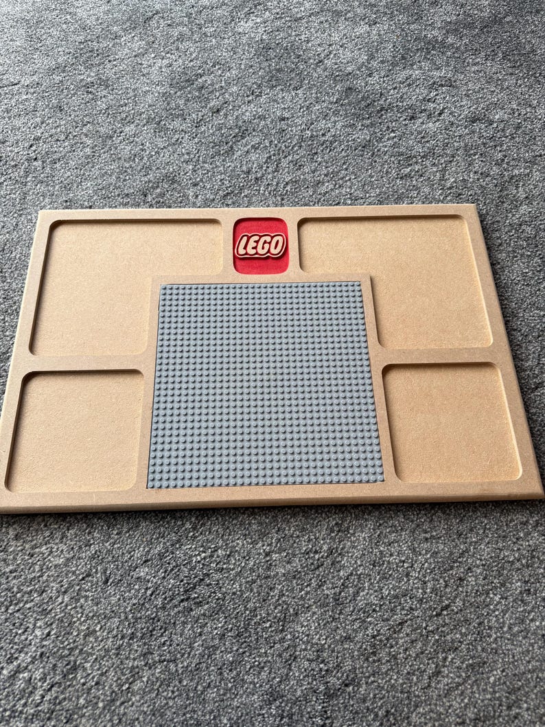 May include: A wooden Lego baseplate with a gray Lego building surface and four compartments. The Lego logo is in a red square at the top. The baseplate is tan.